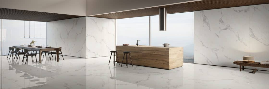 Cozinha com revestimento que reproduz mármore no piso e nas paredes; ilha em madeira com cooktop e banquetas, e uma mesa de jantar.
