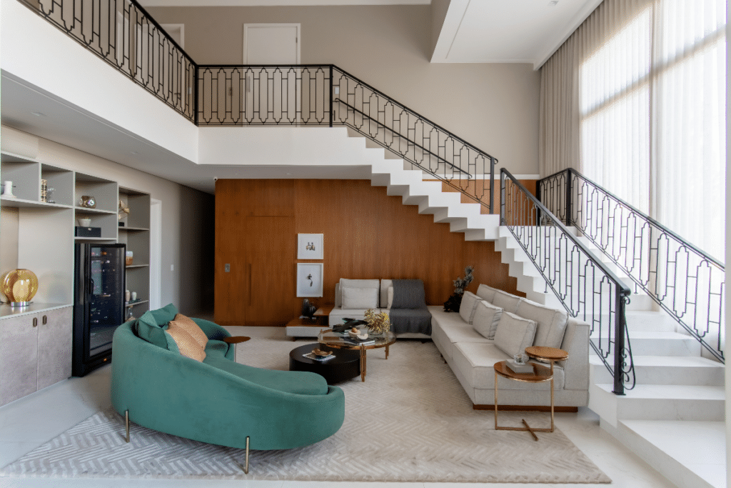Painel em madeira embaixo da escada na sala de estar, com sofá cinza e outro sofá verde. A tonalidade da madeira complementa os tons neutros dos móveis (Projeto: Paiva e Passarini Arquitetura | Imagem: Xavier Neto).