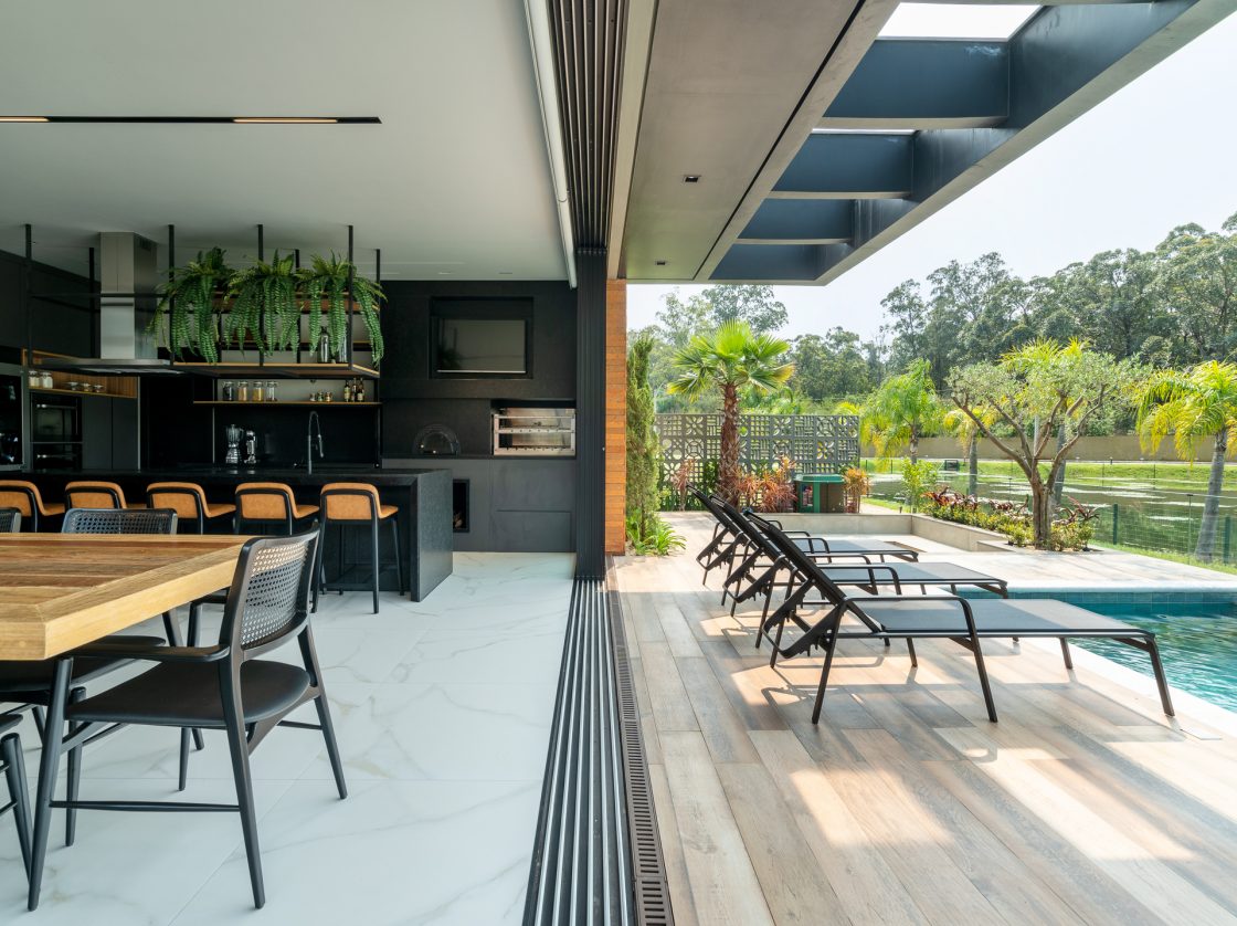 Outdoor Flooring: Discover the Best Options 9 Varanda integrada com espaço gourmet, porcelanato amadeirado e porcelanato marmorizado se encontram, vegetação abundante, piscina e área gourmet em madeira e preto