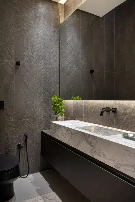 Banheiro com foco de iluminação na parte da pia embaixo do espelho. Uma planta na decoração. Paredes com revestimento em tons cinzas.